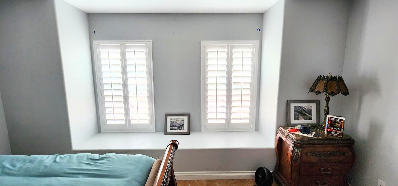 0891a98c-faed-409a-8d09-0cec5e7c0387 Bright white plantation shutters adding elegance and privacy to a cozy bedroom nook with classic decor and natural light.