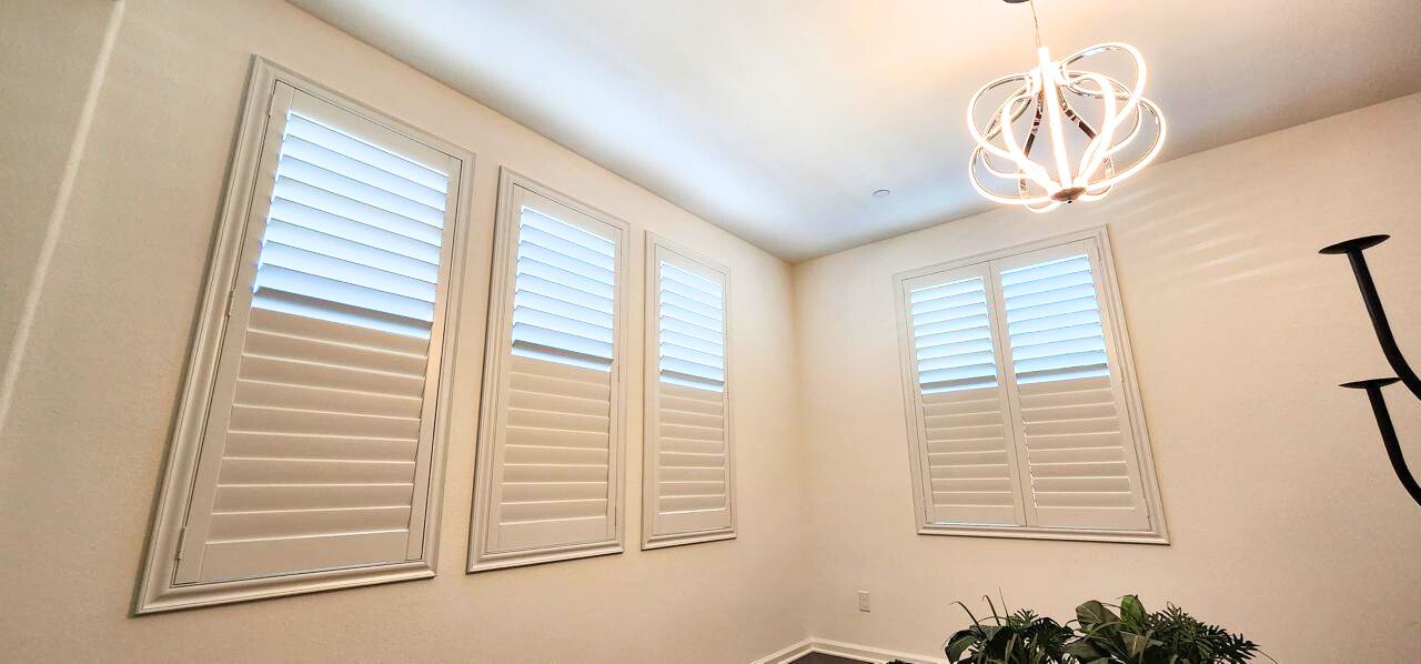 618bd4dd-fb64-4658-b712-af57820a8d29 A room with white walls featuring three tall windows with white shutters and white ceiling lights. There is a white chandelier and some green plants in the corner