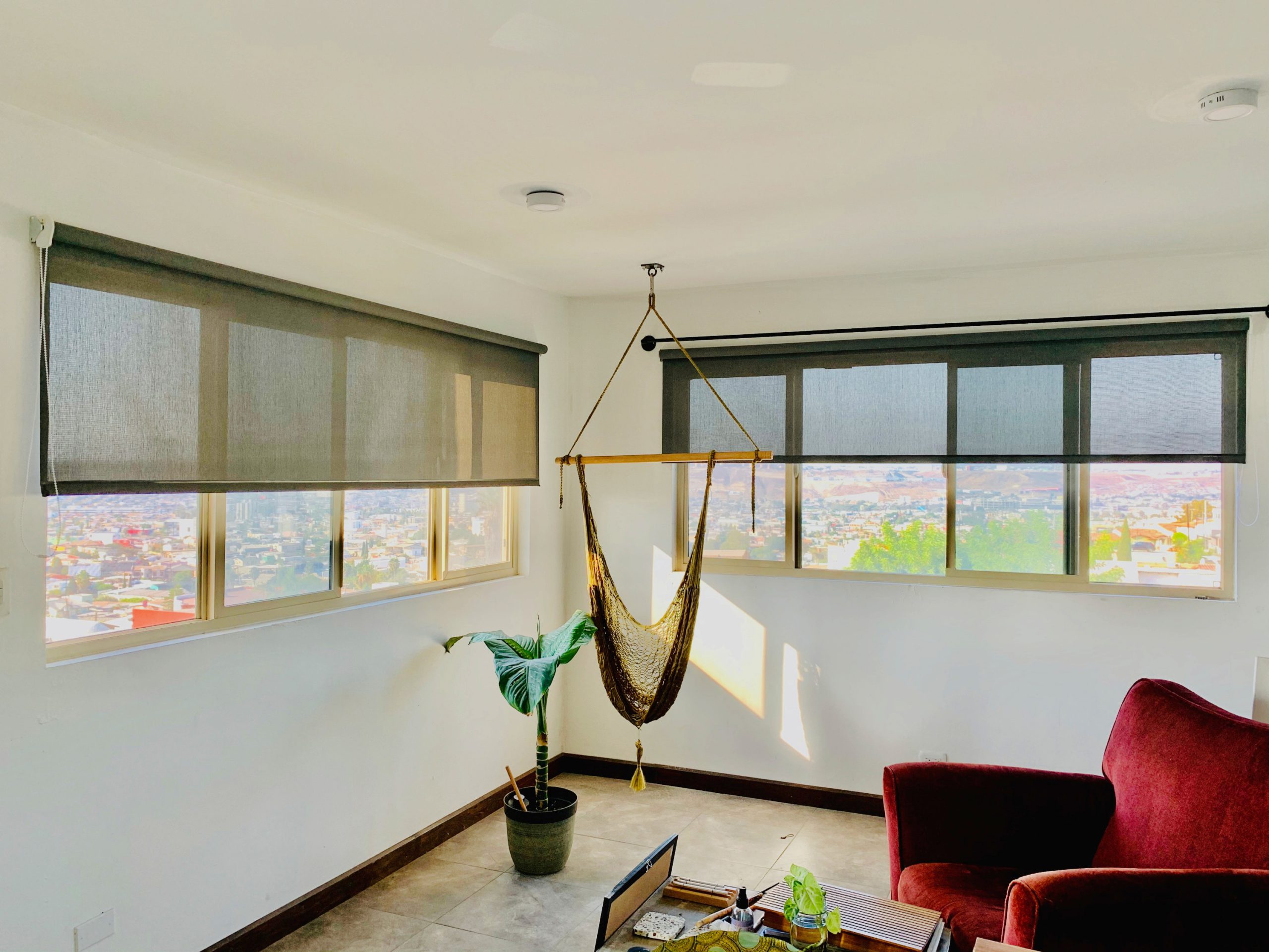 A room with two large windows covered by gray roller shades, a hanging chair, a red armchair, a potted plant, and a small table. The view outside shows a cityscape with hills in the distance.